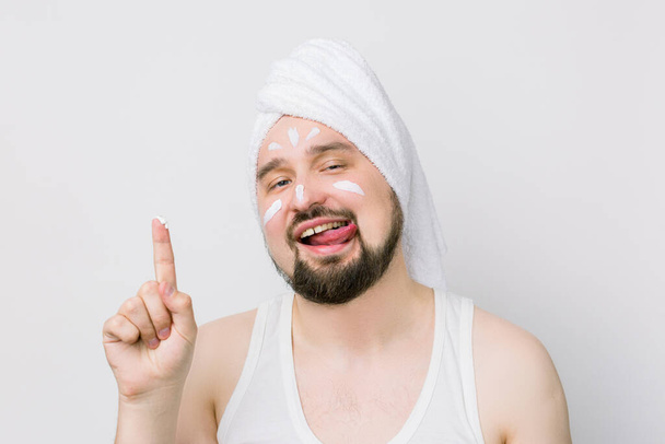 Funny bearded man with white towel on his head showing his finger with face cream on it, smears a cream on his face, smiling and showing his tongue. The concept of male self-care and male cosmetics - Photo, Image
