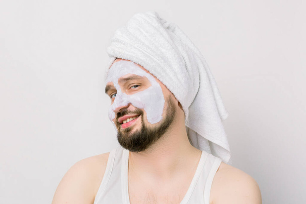 Portrait of a handsome joyful relaxed young bearded man with a facial mud clay mask, with towel on his head, posing with funny emotions on isolated white background. Face and body skin care treatment. - Foto, imagen