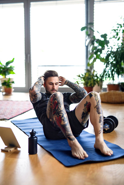 Front view portrait of man with tablet doing workout exercise indoors at home. - Фото, изображение