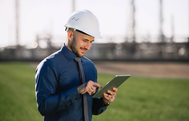 Young engineer with hard hat and tablet standing outdoors by oil refinery. - Foto, Bild