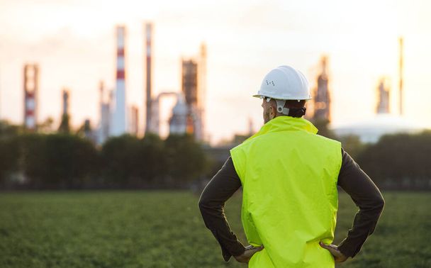 Rear view of young engineer standing outdoors by oil refinery, arms on hips. - Foto, Imagem