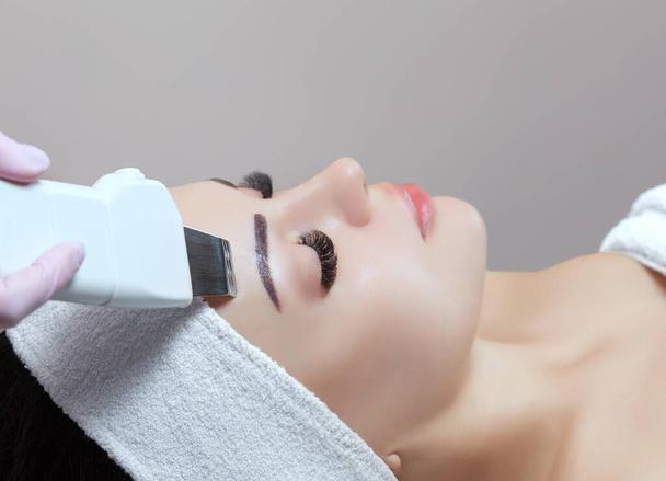 De arts-cosmetoloog maakt van het apparaat een procedure voor ultrasone reiniging van de gezichtshuid van een mooie, jonge vrouw in een schoonheidssalon. Cosmetologie en professionele huidverzorging. - Foto, afbeelding