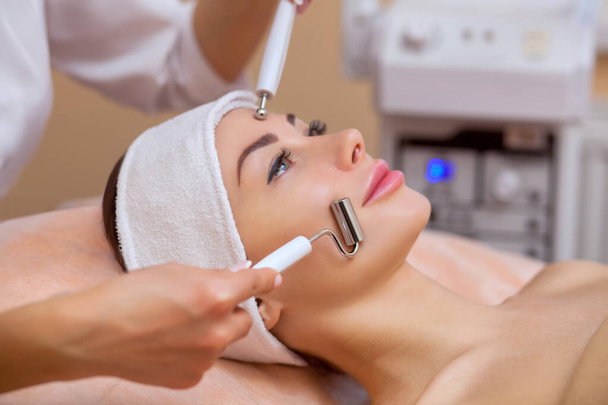 The physician-cosmetologist makes the apparatus a procedure of "galvanic" ultrasound cleaning of the face skin of a beautiful, young woman in a beauty salon. Cosmetology and professional skin care. - Photo, Image