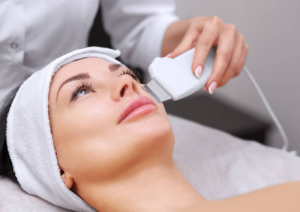 The doctor-cosmetologist makes the apparatus a procedure of ultrasound cleaning of the facial skin of a beautiful, young woman in a beauty salon. Cosmetology and professional skin care. - Photo, Image