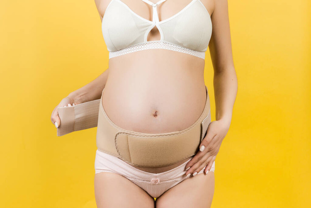 Zugeschnittenes Bild einer schwangeren Frau in Unterwäsche, die einen Stützverband anlegt, um Rückenschmerzen auf gelbem Hintergrund mit Kopierraum zu lindern. Orthopädisches Bauchstützgurt-Konzept. - Foto, Bild