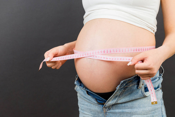 Imagen recortada de la madre embarazada en jeans desabrochados midiendo su vientre embarazada con una cinta métrica en un fondo colorido. medición de centímetros. - Foto, Imagen