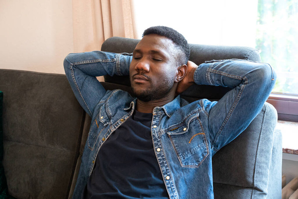 Black man resting on couch with eyes closed and hands behind head. Medium shot.  - Φωτογραφία, εικόνα