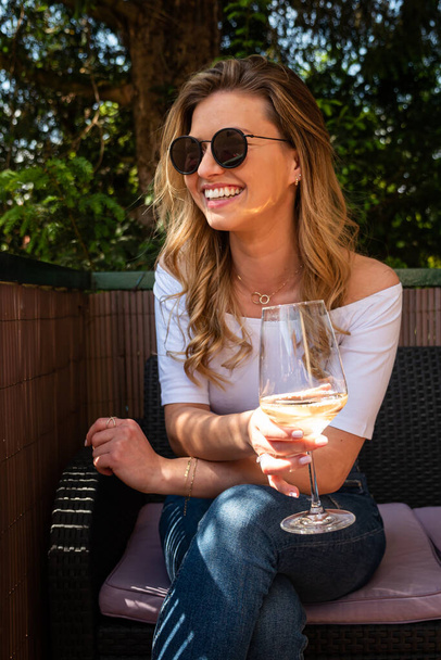 Mulher loira bonita usando óculos de sol e segurando copo de vinho ao ar livre sorrindo. A desviar o olhar. Comprimento de três quartos
.  - Foto, Imagem