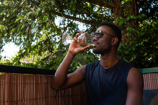 Young black man drinking wine seated outdoors. Medium shit. Side view.  - 写真・画像