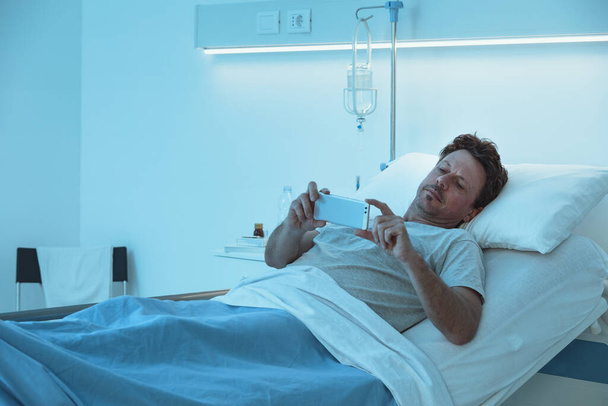 Adult sleepless patient lying in a hospital bed at night, he is watching video with his smartphone - Zdjęcie, obraz