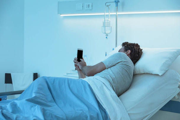 Безсонний стурбований пацієнт лежить в ліжку вночі в лікарні і спілкується зі своїм смартфоном
 - Фото, зображення