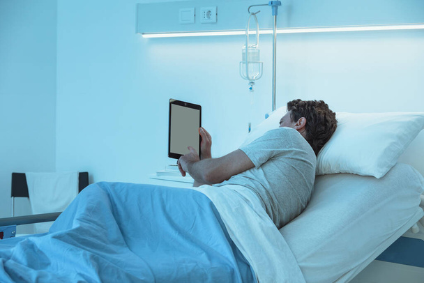 Dorosły bezsenny pacjent leżący w nocy w szpitalnym łóżku; pracuje nad swoim tabletem cyfrowym - Zdjęcie, obraz