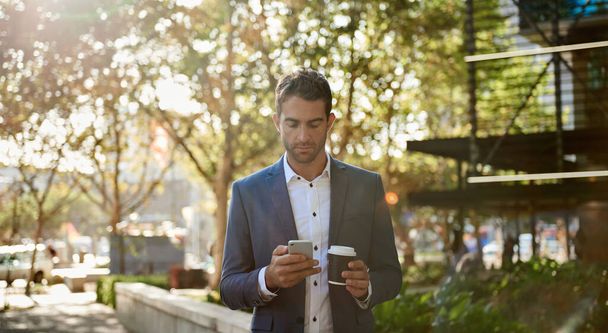 Genç işadamı, şehirde elinde kahve fincanıyla yürürken cep telefonundan bir mesaj okuyor. - Fotoğraf, Görsel