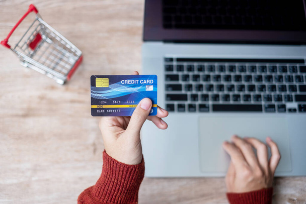 woman wearing sweater holding credit card and using laptop for online shopping while making order at home office. business, lifestyle, technology, ecommerce, digital banking and online payment concept - Photo, Image