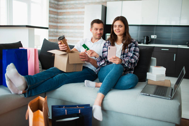 Ein junges Ehepaar kauft online ein. Um sie herum liegt eine Menge Ware in Papierverpackungen, Schachteln und Tüten.. - Foto, Bild