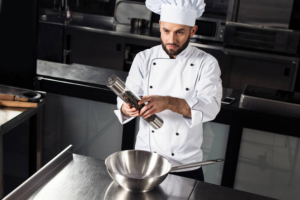 Male chef with wok at kitchen restaurant. Concentrated man with pepperbox. - Photo, Image