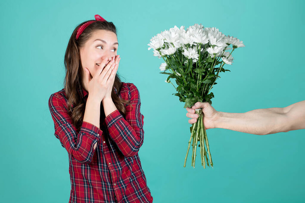 A country girl in a red plaid shirt laughs, covering her mouth and looking at the flowers that a man gives her - a bouquet of white chrysanthemums - Foto, Imagen