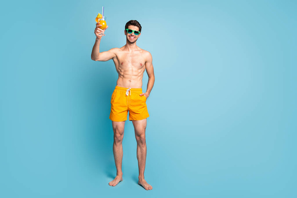 Full length body size photo of cheerful handsome attractive man with hand in yellow shorts pocket standing near ocean isolated pastel blue color background - Foto, immagini