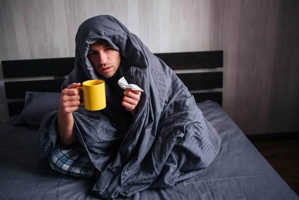 He covered himself with a blanket and holds a napkin and hot tea. The man got sick. Lies in bed and suffers. He has fever, flu, colds and snot. - Фото, изображение
