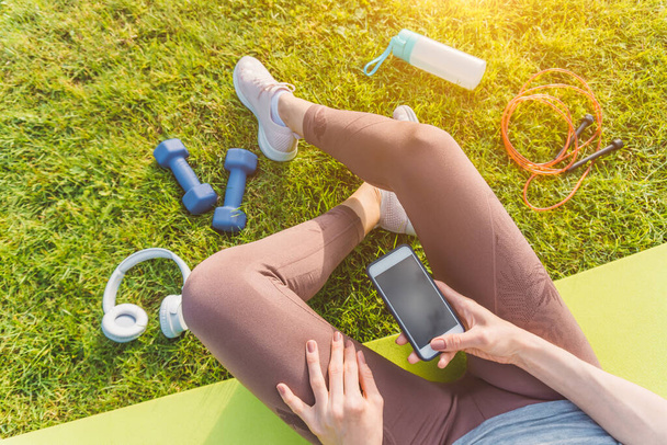 Flat lay picture of a girl sitting on green grass with sports equipment holding smartphone in her hands - Photo, Image