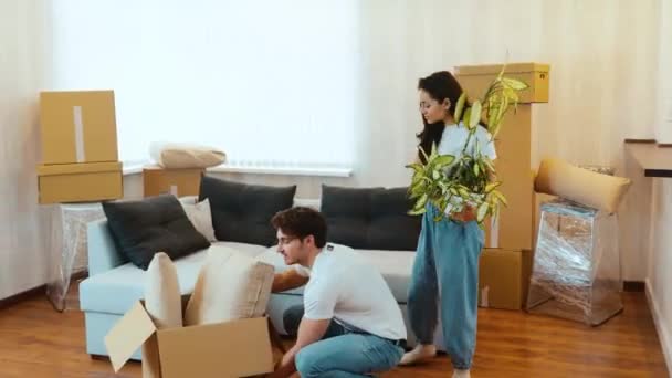 Jong stel verhuist naar nieuw appartement. Een man draagt een doos met kussens erin en legt het op de vloer. Jonge vrouw volgt hem met een plant. Geniet van hun nieuwe appartement. - Video