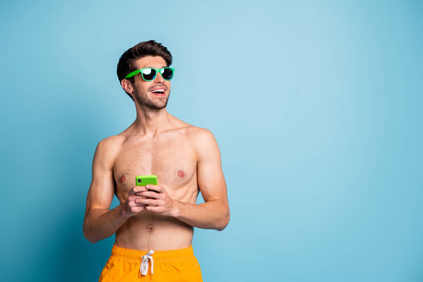 Photo of cheerful positive handsome man looking into empty space with torso naked yellow shorts browsing through telephone in sunglass isolated pastel blue color background - Valokuva, kuva