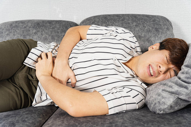 Nuori mies kärsii vatsakivusta makatessaan sohvalla kotona. Terveydenhuollon lääketieteellisen tai jokapäiväisen elämän käsite. - Valokuva, kuva