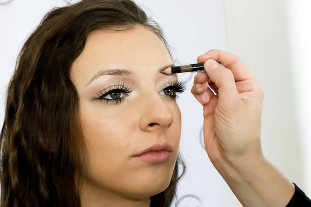 Make-up-Artist arbeitet mit Stift auf Modell Augenbrauen Nahaufnahme des Schminkens auf junge Mädchen, Semi-Profil, professionelles Make-up im Schönheitsstudio - Foto, Bild