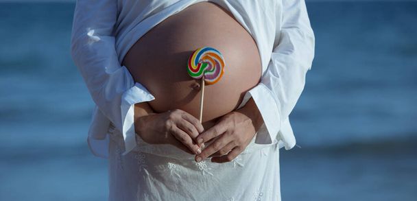 Genç hamile bir kadın sahilde ve elinde büyük renkli bir lolipop tutuyor. - Fotoğraf, Görsel