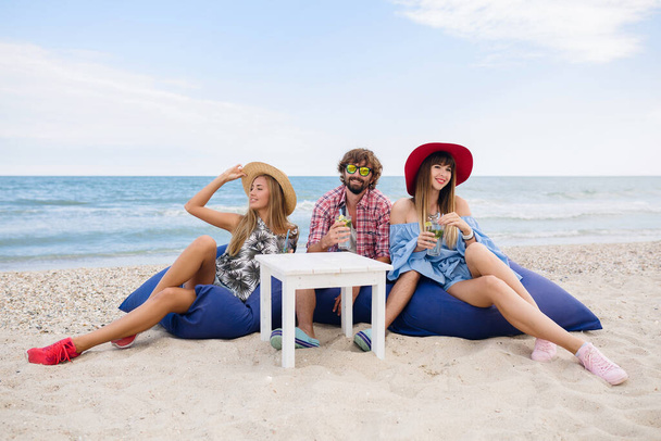 young hipster company of friends on vacation sitting at beach on bean bags, drinking mojito cocktail, happy, smiling, positive, summer style, fashion trend, spending good time together, party mood - Photo, Image