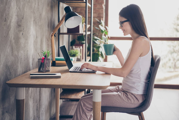 Profile side photo of pretty positive girl agent executive work distance use laptop have partners conference video call hold mug beverage sit desk chair in house indoors - 写真・画像