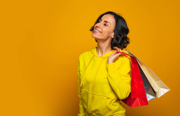 Can't wait to wear new clothes! Half-length photo of a young woman, turned sideways, caring her purchase in colorful bags behind her shoulder and dreaming about her new clothes. - Fotó, kép