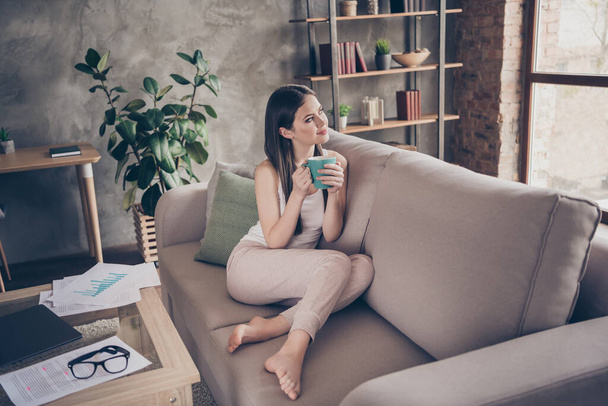 Full length photo of interested positive girl have free time after routine remote work hold beverage look dream dreamy sit divan in house indoors - Фото, изображение