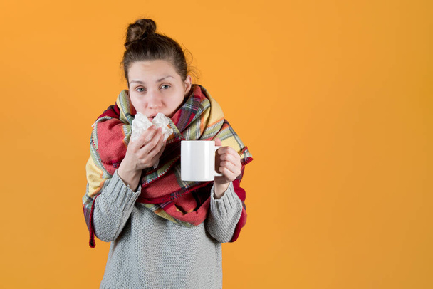 A young woman with a hot drink wrapped in a blanket wipes her nose with a napkin. Isolated on orange yellow background. - Zdjęcie, obraz