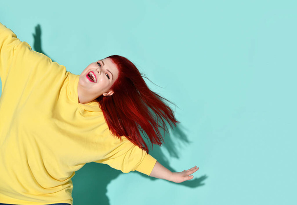 Happy plump female with flowing red hair dancing or jumping with her arms outstretched widely like wings. Cropped shot isolated on blue - Foto, Imagem