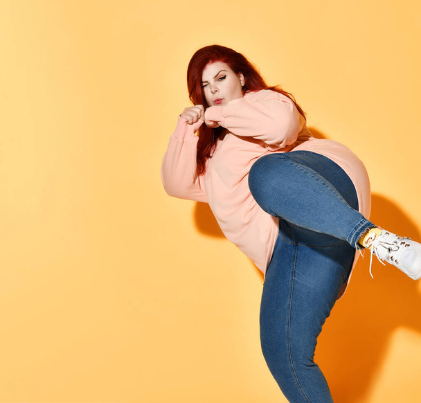 Concentrated fatty young female in jeans and sweater posing in fighting stance preparing to kick. Studio shot isolated on orange, copy space - Fotografie, Obrázek