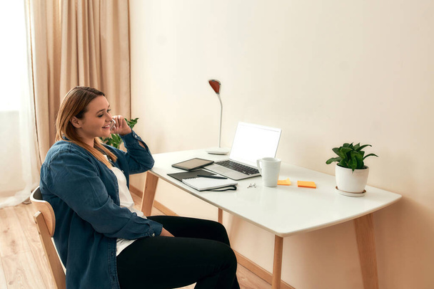 Good talk. Young happy female freelancer in casual clothes sitting at her creative workplace at home, talking on phone with client and smiling - 写真・画像
