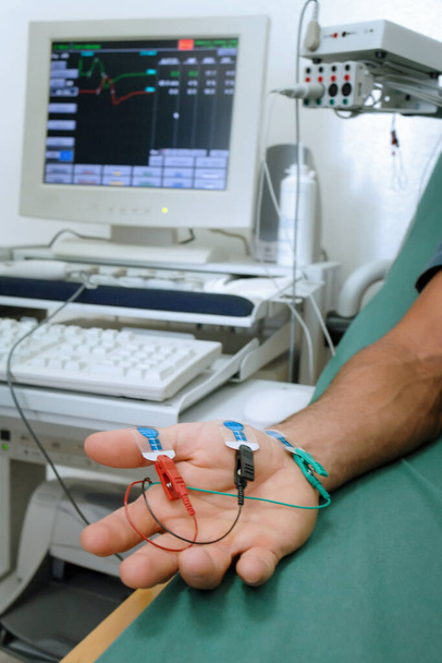 Εργαστηριακές δοκιμές με ηλεκτρόδια στο χέρι ενός ασθενούς που βρίσκεται στο κρεβάτι. Στο παρασκήνιο, ηλεκτρονικός εξοπλισμός για την παρακολούθηση των αποτελεσμάτων των δοκιμών. - Φωτογραφία, εικόνα