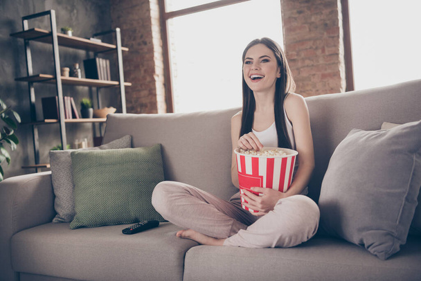 Full length photo of candid girl sit divan legs crossed watch funny movie hold box eat pop corn enjoy free time in house indoors living room - Fotó, kép