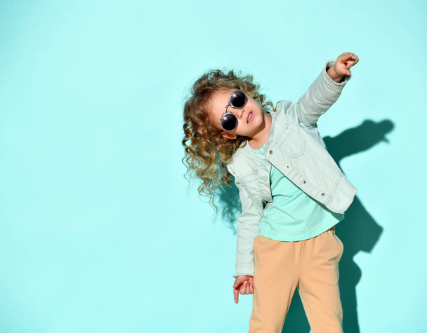 Cute little curly girl with black non-transparent glasses on face stands raising one hand pointing two fingers up. Studio shot isolated on turquoise - Fotografie, Obrázek