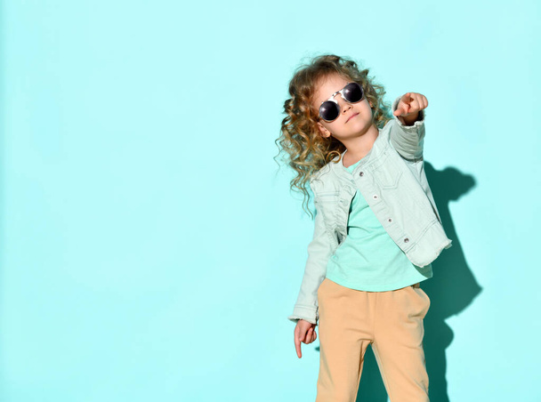 Cute little curly girl with black non-transparent glasses on face stands raising one hand pointing two fingers up. Studio shot isolated on turquoise - Fotografie, Obrázek