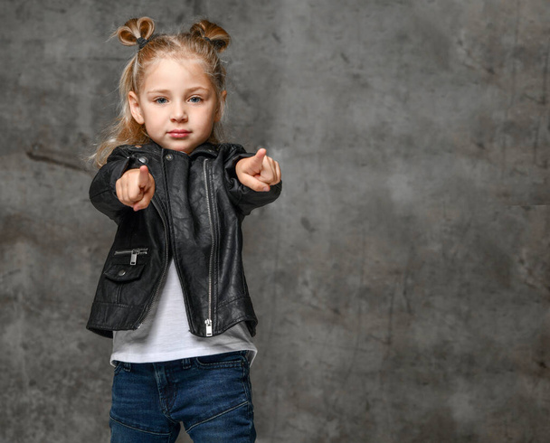 Little cute blond girl in stylish rock style black leather jacket, jeans and sneakers standing and pointing at camera with fingers - Valokuva, kuva