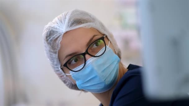 Μια νεαρή γιατρός με γυαλιά κοιτάζει την οθόνη κατά τη διάρκεια της επέμβασης. Κοίτα από κοντά. Εργαζόμενοι στον τομέα της υγείας στην πανδημία του Coronavirus Covid19. - Πλάνα, βίντεο