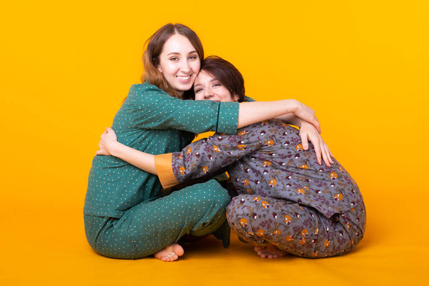 Portré két gyönyörű fiatal lány visel színes pizsamát szórakozás közben ottalvós izolált sárga háttér. Pizsama party és tyúk-party koncepció - Fotó, kép