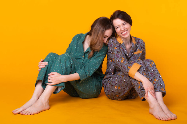 Portrait of two beautiful young girls wearing colorful pyjamas having fun during sleepover isolated over yellow background. Pajama party and hen-party concept - Valokuva, kuva
