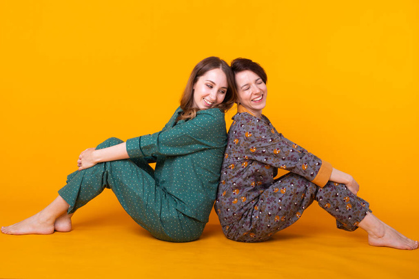 Portret van twee mooie jonge meisjes die kleurrijke pyjama 's dragen en plezier hebben tijdens een slaapfeestje op een gele achtergrond. Pajama partij- en kippenpartijconcept - Foto, afbeelding