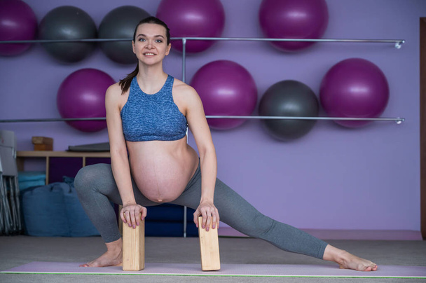 Pregnant flexible European woman is engaged in fitness. The expectant mother in the last trimester of pregnancy practices yoga with the help of yoga blocks. - Fotó, kép