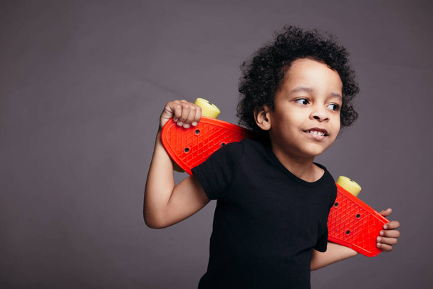Κοντινό πλάνο πορτρέτο του μικρού μαύρου αγοριού σε ένα μαύρο T-shirt κρατώντας κόκκινο skateboard πίσω από την πλάτη του και χαμογελώντας. - Φωτογραφία, εικόνα