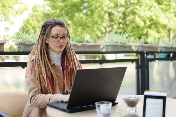 Γοητευτική γυναίκα επιχειρηματίας σε γυαλιά ηλίου με μακριά dreadlocks casual ντυμένος σχεδιάζει μια διαδικασία εργασίας χρησιμοποιώντας εφαρμογή στη συσκευή laptop, ενώ κάθεται στο σύγχρονο δρόμο καφέ σε εξωτερικούς χώρους - Φωτογραφία, εικόνα