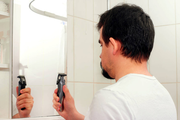 Adult man with disheveled hair standing in front of mirror and looking at hair trimmer (clipper) and thinking about shaving off his hair or beard. Midlife crisis, adult man life, male problems and masculinity problems concept - Фото, зображення